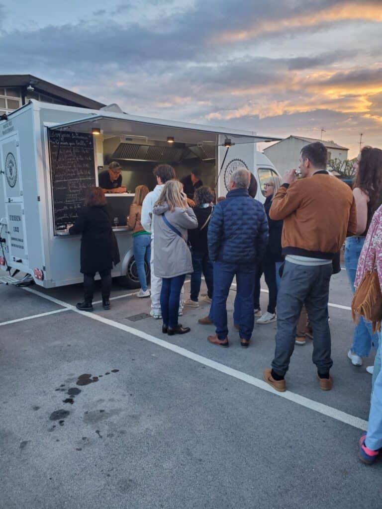 Clientèle faisant la queue devant un food truck gris au crépuscule ; menu lisible : "Bonjour bienvenue au camion qui grille".