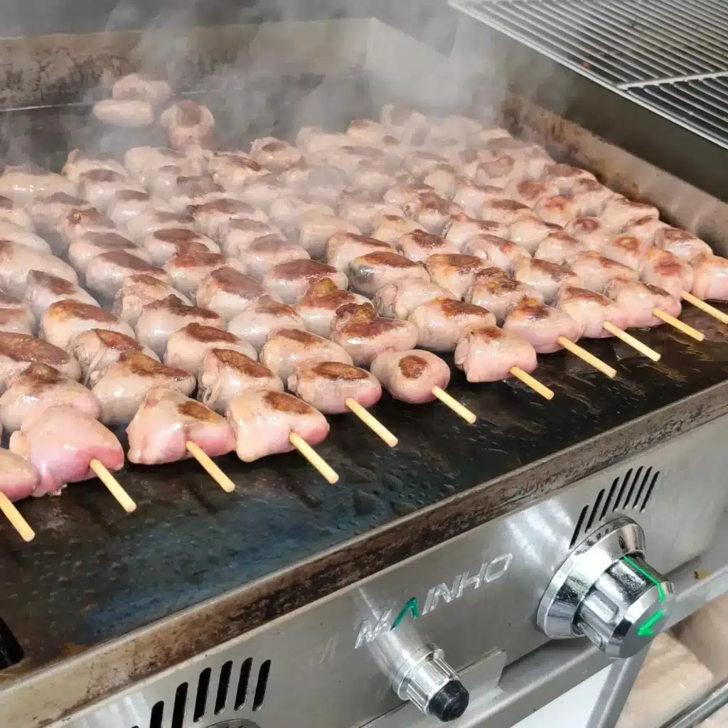 Brochettes de saucisses ou de viande rosée grillant sur une plancha professionnelle MANINHO, dégageant de la vapeur.