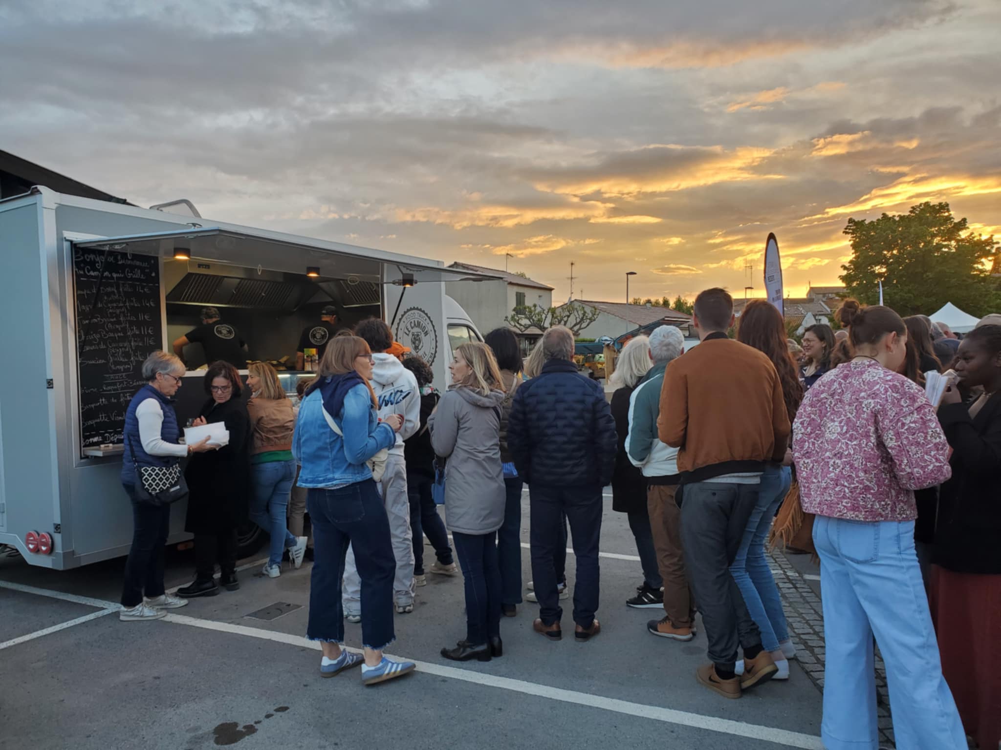 Foule attendant des plats devant un food truck « LE CAMION » au coucher du soleil. Le menu indique des prix comme 14€ et 11€.