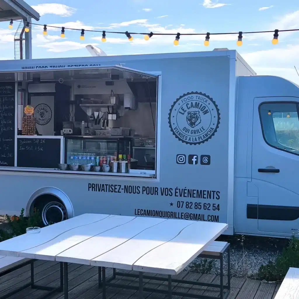 Food truck 'Le Camion Qui Grille' gris avec menu sur ardoise, tables blanches extérieures et guirlande lumineuse.