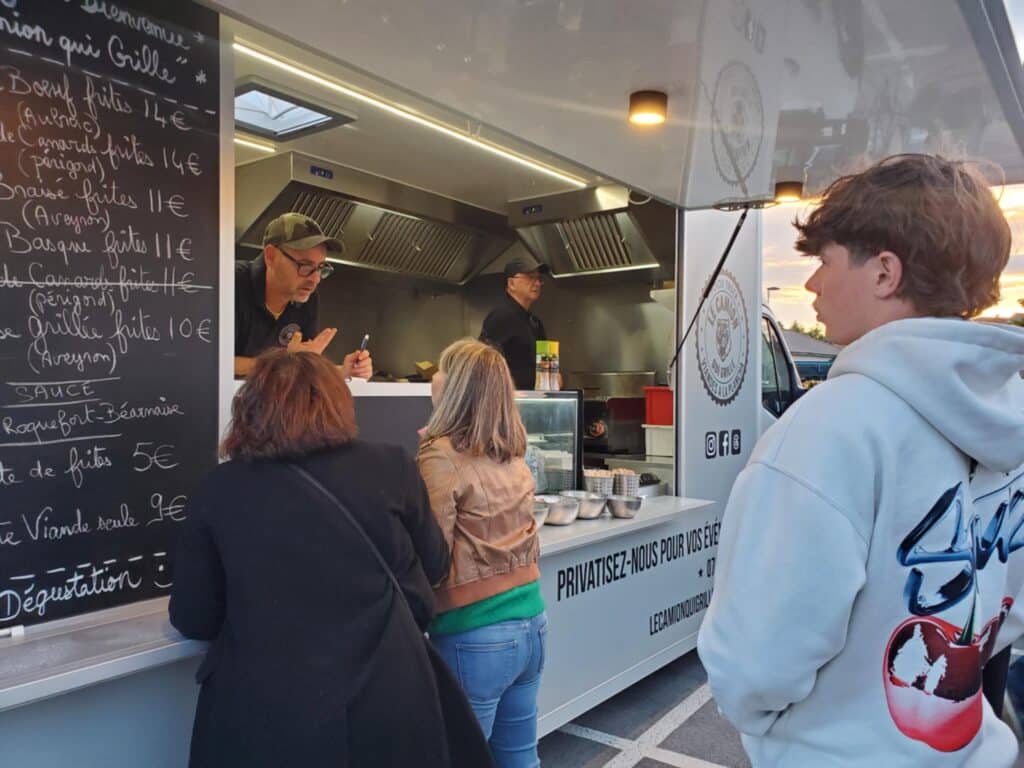 Clients commandant dans un food truck 'Le Camion Qui Grille' avec un menu affiché : Boeuf frites 14€, Canard frites 14€, Sauces Roquefort-Béarnaise 5€.