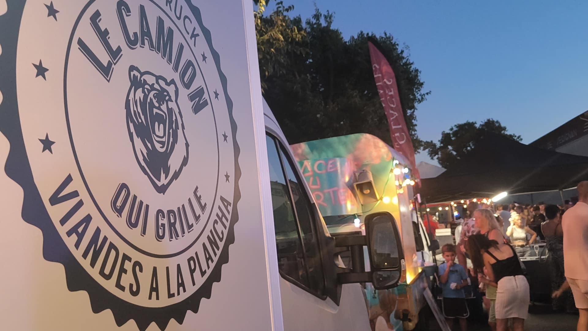 Panneau du food truck "LE CAMION QUI GRILLE VIANDES A LA PLANCHA" avec logo d'ours devant une foule en soirée.