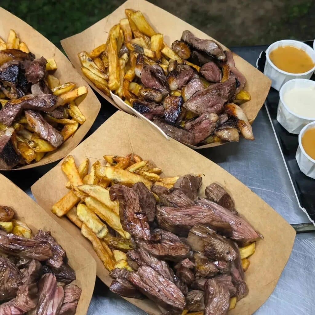 Steak Frites en Barquette : Repas de Rue Savoureux Quatre barquettes de frites dorées garnies de tranches de steak grillé, accompagnées de coupelles de sauces jaune et blanche.